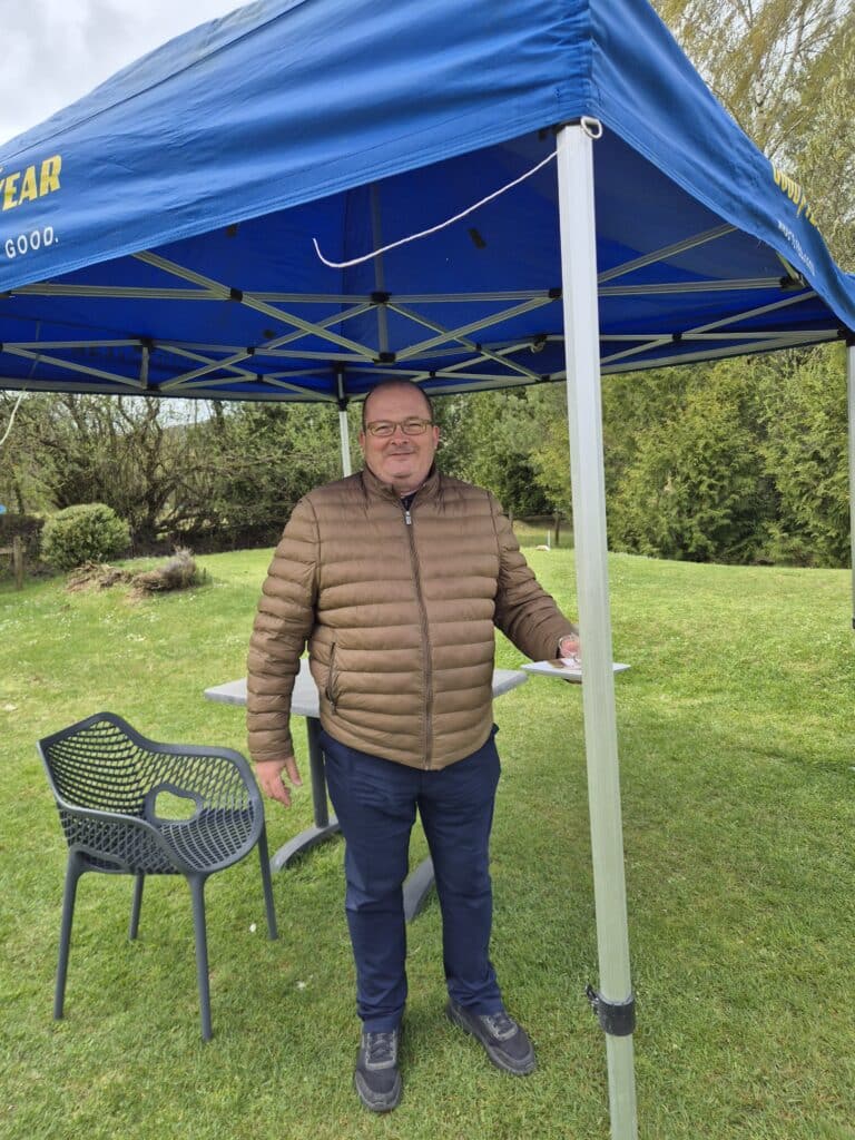 Homme souriant sous un abri de jardin bleu, portant une veste en duvet marron. Il se tient debout sur une pelouse verte, à côté d'une table et d'une chaise noire design. L'environnement naturel est verdoyant, avec des arbres en arrière-plan, suggérant une journée agréable en plein air. Cette scène pourrait évoquer des moments de détente ou des activités en extérieur.