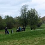 Trois golfeurs se préparent sur un parcours de golf verdoyant, entourés d'arbres et de paysages naturels. L'image montre un homme en train de ranger son sac de golf, tandis que deux autres discutent près de leurs équipements. Le ciel est nuageux, suggérant une journée fraîche, idéale pour le golf. Ce cadre paisible met en avant l'attrait des activités de plein air et le plaisir du golf en harmonie avec la nature.