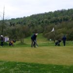 Un terrain de golf verdoyant avec des golfeurs en action, dont un joueur effectue un putt sur le green. En arrière-plan, des caddies sont visibles, tandis que des collines boisées ajoutent une touche naturelle au paysage. L'image illustre une journée de golf agréable, propice à la pratique de ce sport en plein air.