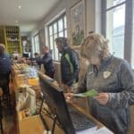 Scène d'un accueil dans un club de golf, montrant un comptoir avec des employés en train de servir des clients. Une femme consulte des documents près d'un ordinateur, tandis que plusieurs personnes, habillées en tenue de golf, attendent au bar. Des balles de golf et divers articles sont visibles sur le comptoir. L'ambiance est conviviale et sportive, typique d'un club de golf.