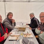 Quatre hommes assis autour d'une table à l'intérieur d'une tente. Ils partagent un moment convivial après un repas, avec des assiettes partiellement vides et des verres à vin sur la table. L'ambiance est détendue, et les participants portent des vêtements décontractés adaptés à la saison. Le décor est simple, avec un fond blanc qui met en valeur les convives. Cette image illustre un moment de camaraderie et de convivialité, idéal pour des