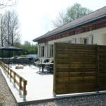 Vue d'un espace extérieur aménagé avec une terrasse en béton, entourée de panneaux en bois. À gauche, un abri de jardin avec une table et des chaises, idéal pour des repas en plein air. L'environnement verdoyant, avec des arbres en arrière-plan, offre une atmosphère paisible. Cet aménagement extérieur est parfait pour des moments de détente ou des activités en famille.