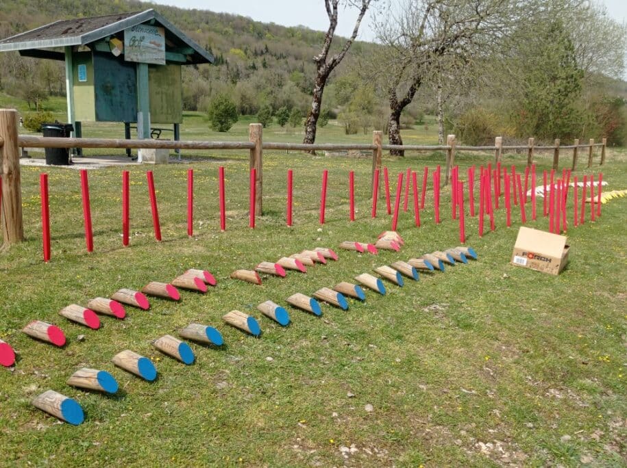 Image d'un terrain de golf avec des obstacles colorés. Au premier plan, des poteaux rouges et des éléments en bois peint en bleu et rose sont disposés sur l'herbe. En arrière-plan, un abri en bois avec un panneau d'information sur le golf. L'environnement est verdoyant, avec des arbres et une clôture en bois. Cette image illustre un espace de loisirs en plein air, idéal pour des activités familiales et sportives.