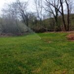 Arrosage automatique d'une pelouse verdoyante avec un système de sprinkleurs. En arrière-plan, des arbres dénudés et des broussailles. Cette image illustre l'entretien d'un jardin, mettant en avant l'importance de l'irrigation pour une croissance saine de l'herbe. Les couleurs vives du gazon soulignent l'efficacité de l'arrosage.