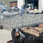 Barbecue convivial en plein air avec des viandes grillées, incluant des saucisses et des morceaux de viande marinée. En arrière-plan, des personnes discutent et se détendent autour d'une ambiance amicale. Le décor inclut des murs en gabion et des tables, créant un cadre agréable pour profiter d'un repas en plein air. Cette image illustre un moment de partage et de gastronomie en extérieur, parfait pour des événements sociaux ou des rassemblements familiaux.