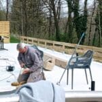Un homme travaille sur un chantier en extérieur, occupé à utiliser une perceuse sur une surface en béton. En arrière-plan, on aperçoit une table en métal et des chaises en plastique. Une clôture en bois délimite l'espace, tandis que des arbres feuillus apportent une touche naturelle à l'environnement. Des outils et des matériaux sont visibles, témoignant d'un projet de construction ou de rénovation en cours. Cette image illustre le travail manuel et l'aménagement de l