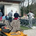 Groupe de personnes réunies à l'extérieur d'un bâtiment, profitant d'un moment convivial autour d'une table. L'environnement est paisible, avec des arbres en arrière-plan et une pelouse bien entretenue. Un véhicule de jardinage est garé à proximité, suggérant une activité de plein air. Les participants semblent discuter et partager des repas, illustrant une ambiance de convivialité et de détente.