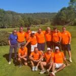 Groupe d'hommes en polo orange posant sur un terrain de golf verdoyant par une belle journée ensoleillée. Ils affichent des sourires et semblent profiter d'un moment convivial ensemble. En arrière-plan, on aperçoit d'autres golfeurs et un paysage boisé, ajoutant à l'atmosphère de détente et de camaraderie. Cette image évoque l'esprit du golf et les rencontres amicales entre passionnés.
