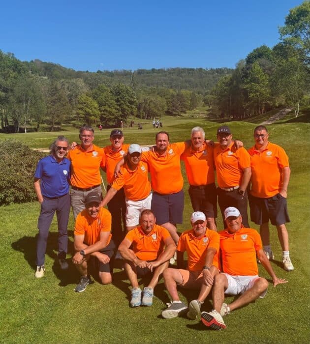 Groupe d'hommes en polo orange posant sur un terrain de golf verdoyant par une belle journée ensoleillée. Ils affichent des sourires et semblent profiter d'un moment convivial ensemble. En arrière-plan, on aperçoit d'autres golfeurs et un paysage boisé, ajoutant à l'atmosphère de détente et de camaraderie. Cette image évoque l'esprit du golf et les rencontres amicales entre passionnés.