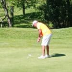 Un golfeur en tenue décontractée, avec un polo orange et un short blanc, se concentre sur un putt sur un green parfaitement entretenu. La scène est entourée de verdure, avec des arbres en arrière-plan, créant une atmosphère paisible et agréable pour le golf. Cette image illustre l'esprit du jeu et la passion pour ce sport en plein air.