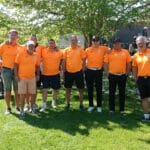Équipe de golf vêtue de polos orange, réunie sur un terrain verdoyant lors d'un événement sportif. Les membres affichent un esprit d'équipe et de camaraderie, avec des accessoires de golf visibles en arrière-plan. L'image illustre une journée ensoleillée, idéale pour la pratique du golf, et met en avant l'importance de l'esprit communautaire dans ce sport.