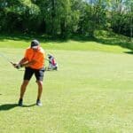 Un golfeur en tenue orange effectue un swing sur un parcours verdoyant. En arrière-plan, un chariot de golf est stationné, tandis que des arbres bordent le terrain. Cette image illustre l'expérience de jeu en plein air, mettant en avant la passion pour le golf et les équipements associés. L'environnement naturel et ensoleillé crée une ambiance idéale pour une journée de golf.