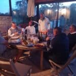 Groupe d'amis réunis autour d'une table dans un espace extérieur, profitant d'un repas convivial en soirée. Les participants, de différents âges, partagent des plats variés et des bouteilles de vin, tandis que la lumière douce du crépuscule crée une ambiance chaleureuse. L'image évoque des moments de camaraderie et de détente, illustrant l'importance des repas partagés dans les relations sociales.