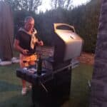 Un homme prépare un barbecue en extérieur, sous un ciel crépusculaire. Il porte un t-shirt noir et un pull noué autour de son cou. À côté de lui, une bouteille de vin rouge est posée sur le barbecue. L'environnement est verdoyant, avec des murs en pierre et un espace gazonné, créant une ambiance conviviale et chaleureuse pour une soirée d'été.