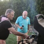 Deux hommes préparent un barbecue en extérieur, profitant d'une belle journée. L'un d'eux, vêtu d'un t-shirt noir, manipule la grille du barbecue où cuisent des morceaux de viande. L'autre, en t-shirt bleu, tient un verre de vin rouge et observe attentivement la cuisson. En arrière-plan, un jardin verdoyant crée une ambiance conviviale. Cette scène évoque des moments de partage et de convivialité autour d'un repas en plein air.