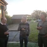 Trois hommes discutent en extérieur lors d'une soirée conviviale, chacun tenant un verre à la main. L'un d'eux porte un pull noir avec un col blanc, tandis qu'un autre arbore un pull à rayures. Le troisième homme est vêtu d'une chemise grise. En arrière-plan, un paysage verdoyant est visible, avec un abri en bois et des arbres. La lumière du coucher de soleil crée une ambiance chaleureuse et détendue, idéale pour des