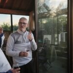 Groupe d'hommes en discussion sur une terrasse en bois, au crépuscule. Deux hommes portent des vêtements de sport, l'un avec une chemise blanche et l'autre en noir, tandis qu'un troisième, en pull gris, tient un verre de vin. En arrière-plan, on aperçoit des arbres et une lumière douce du soleil couchant à travers une grande fenêtre. Cette scène évoque une ambiance conviviale et détendue, idéale pour des moments de partage et de convivialité.