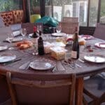 Table dressée pour un repas convivial en extérieur, avec des assiettes décoratives, des verres à vin et plusieurs bouteilles de vin. On y trouve également des croissants, des plats de nourriture et des couverts bien disposés sur une table ronde en bois. L'ambiance chaleureuse est accentuée par un cadre naturel, idéal pour un dîner entre amis ou en famille.