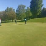 Trois golfeurs sur un green ensoleillé, concentrés sur un putt. L'un des joueurs s'apprête à frapper la balle, tandis que les autres observent attentivement. L'environnement verdoyant est agrémenté d'arbres et d'une pelouse bien entretenue, typique des parcours de golf. Cette scène illustre l'esprit convivial et compétitif du golf en plein air.