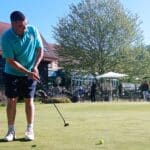 Un golfeur en action sur le green, concentré sur son putt. L'arrière-plan montre une terrasse animée d'un club de golf, entourée d'arbres verdoyants sous un ciel clair. Des joueurs et des spectateurs profitent de la journée ensoleillée. Cette scène illustre l'ambiance conviviale et détendue d'une journée de golf.