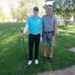 Deux golfeurs posent sur un parcours verdoyant, l'un portant une tenue de golf bleue et l'autre en polo gris. Ils se tiennent côte à côte, souriants, avec un club de golf dans la main du premier. En arrière-plan, on aperçoit des arbres et un chemin, indiquant une belle journée ensoleillée propice au golf. Cette image illustre l'esprit de camaraderie et le plaisir de jouer au golf en plein air.