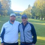 Deux golfeurs posent sur un parcours verdoyant, entourés d'arbres majestueux. L'image capture l'esprit convivial du golf, avec des joueurs vêtus de tenues sportives adaptées. Le soleil brille, créant une atmosphère agréable pour une journée de golf. Cette scène illustre parfaitement les moments de partage et de détente sur le parcours.