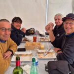 Quatre personnes souriantes autour d'une table, profitant d'un repas convivial. L'environnement est décontracté, avec des assiettes vides et des verres à moitié pleins. On aperçoit des bouteilles de vin et d'eau, ainsi qu'un assortiment de biscuits. Cette image illustre un moment de partage et de gastronomie, parfait pour des articles sur la convivialité en groupe ou sur les repas en plein air.