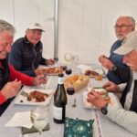 Quatre hommes partagent un repas convivial sous une tente, chacun savourant un plat appétissant. Ils sont entourés de verres de vin et de pain, créant une atmosphère chaleureuse et amicale. Les participants portent des vestes et des casquettes, adaptés à un cadre décontracté. Cette scène évoque des moments de camaraderie et de partage autour de la nourriture.