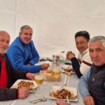 Quatre hommes profitent d'un repas convivial sous une tente. Ils sont assis autour d'une table, chacun avec une assiette de nourriture et un verre de bière. L'ambiance décontractée reflète un moment de partage et de camaraderie. Les participants portent des vêtements adaptés à la saison, et la décoration de la table est simple mais accueillante. Cette image illustre l'importance des repas en groupe et des échanges amicaux.