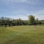Un groupe de golfeurs sur un green verdoyant par une journée ensoleillée. Au premier plan, une golfeuse s'apprête à putter près du drapeau, tandis que d'autres joueurs analysent le terrain. En arrière-plan, des arbres feuillus et un ciel bleu clair complètent le paysage paisible d'un parcours de golf. Cette scène illustre la convivialité et la stratégie du golf, attirant les amateurs de ce sport en plein air.