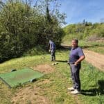 Deux hommes se préparent à jouer au golf sur un parcours verdoyant. L'un d'eux se tient debout près d'un tee, arborant une tenue de golf décontractée, tandis que l'autre s'apprête à frapper la balle. Le paysage environnant est composé d'arbres feuillus et d'un ciel dégagé, créant une atmosphère agréable pour la pratique du golf. Cette image illustre une journée en plein air dédiée au sport et à la convivialité