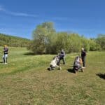 Groupe de golfeurs sur un parcours verdoyant par une belle journée ensoleillée. Trois hommes se préparent à jouer, avec des sacs de golf à roulettes à leurs côtés. L'environnement est calme, entouré d'arbres feuillus et d'un ciel bleu dégagé. Cette scène illustre parfaitement l'esprit du golf en plein air, favorisant la détente et le plaisir du jeu. Idéal pour les amateurs de golf cherchant à profiter de la nature tout en prati