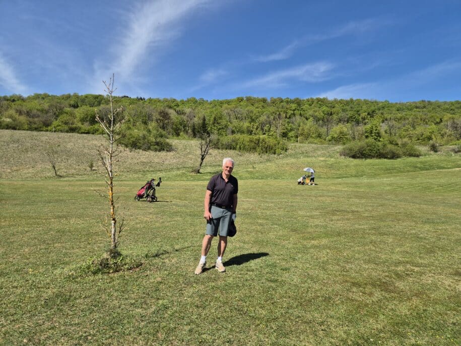 Homme sur un terrain de golf verdoyant, avec un ciel bleu et des nuages épars. En arrière-plan, des golfeurs avec des chariots de golf et un parapluie. Le paysage présente des collines boisées et une pelouse bien entretenue, idéale pour les activités de plein air. Cette scène évoque un moment de détente et de loisir dans un cadre naturel.