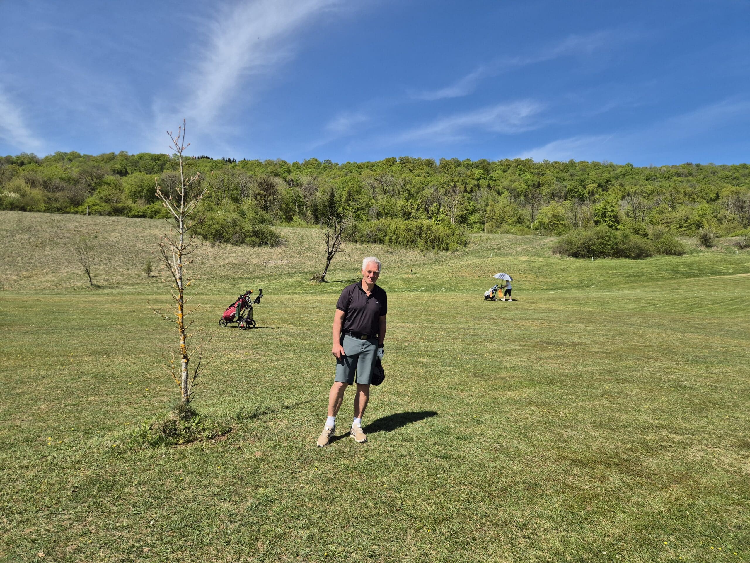 Homme sur un terrain de golf verdoyant, avec un ciel bleu et des nuages épars. En arrière-plan, des golfeurs avec des chariots de golf et un parapluie. Le paysage présente des collines boisées et une pelouse bien entretenue, idéale pour les activités de plein air. Cette scène évoque un moment de détente et de loisir dans un cadre naturel.