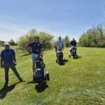 Quatre golfeurs profitent d'une belle journée ensoleillée sur un parcours de golf verdoyant. Ils se déplacent avec leurs chariots de golf, entourés d'arbres en fleurs et d'un ciel bleu clair. L'image illustre l'esprit convivial et détendu du golf, parfait pour les amateurs de ce sport en plein air.