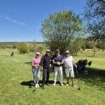 Quatre golfeurs posent sur un parcours verdoyant sous un ciel bleu dégagé. Ils tiennent des clubs de golf et affichent des sourires, prêts à profiter d'une journée de golf. En arrière-plan, on aperçoit d'autres golfeurs et des arbres, ajoutant à l'ambiance paisible d'un club de golf. Cette image illustre parfaitement les moments de convivialité et de détente que l'on peut vivre sur le green.