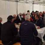 Repas convivial sous un chapiteau, où un groupe d'hommes discute et partage un moment ensemble. Les tables sont dressées avec des assiettes de nourriture et des verres de bière. L'ambiance est chaleureuse, illustrant la camaraderie et le plaisir de se retrouver. Cette image capture l'esprit de convivialité lors d'un événement en plein air, idéal pour promouvoir des activités sociales et des rassemblements communautaires.
