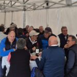 Groupe d'hommes réunis autour d'une table sous un chapiteau, partageant un repas convivial. L'ambiance est détendue, avec des discussions animées et des rires. Plusieurs bouteilles de vin sont visibles sur la table, soulignant l'aspect festif de l'événement. Les participants portent des vêtements décontractés adaptés à la saison, créant une atmosphère chaleureuse et amicale. Cette image illustre un moment de camaraderie lors d'un rassemble