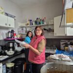 Deux femmes souriantes se trouvent dans une cuisine animée. L'une d'elles, portant un gilet rose, tient des assiettes tandis que l'autre, en arrière-plan, regarde avec un sourire. La cuisine est bien équipée, avec divers appareils électroménagers visibles, tels qu'un robot culinaire et une cafetière. Des plats de riz sont disposés sur le plan de travail, témoignant d'une préparation culinaire en cours. L'espace est organisé, avec des