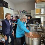 Groupe de bénévoles en cuisine, préparant un repas dans une grande casserole. Une femme souriante en sweat bleu remue la préparation, tandis que d'autres s'affairent en arrière-plan. L'espace de cuisine est bien équipé, avec des ustensiles et des ingrédients visibles. Cette scène illustre la convivialité et la solidarité dans un environnement communautaire.