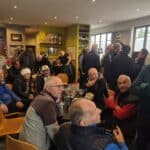 Groupe de personnes réunies dans un café convivial, probablement après une activité sportive. L'ambiance est animée avec des échanges entre amis. On peut apercevoir des coupes et des trophées sur les murs, indiquant un lieu de rencontre pour des passionnés de sport. Les participants semblent apprécier un moment de détente, avec des boissons à leur table. Cette scène illustre la camaraderie et le plaisir de se retrouver autour d'une passion commune.