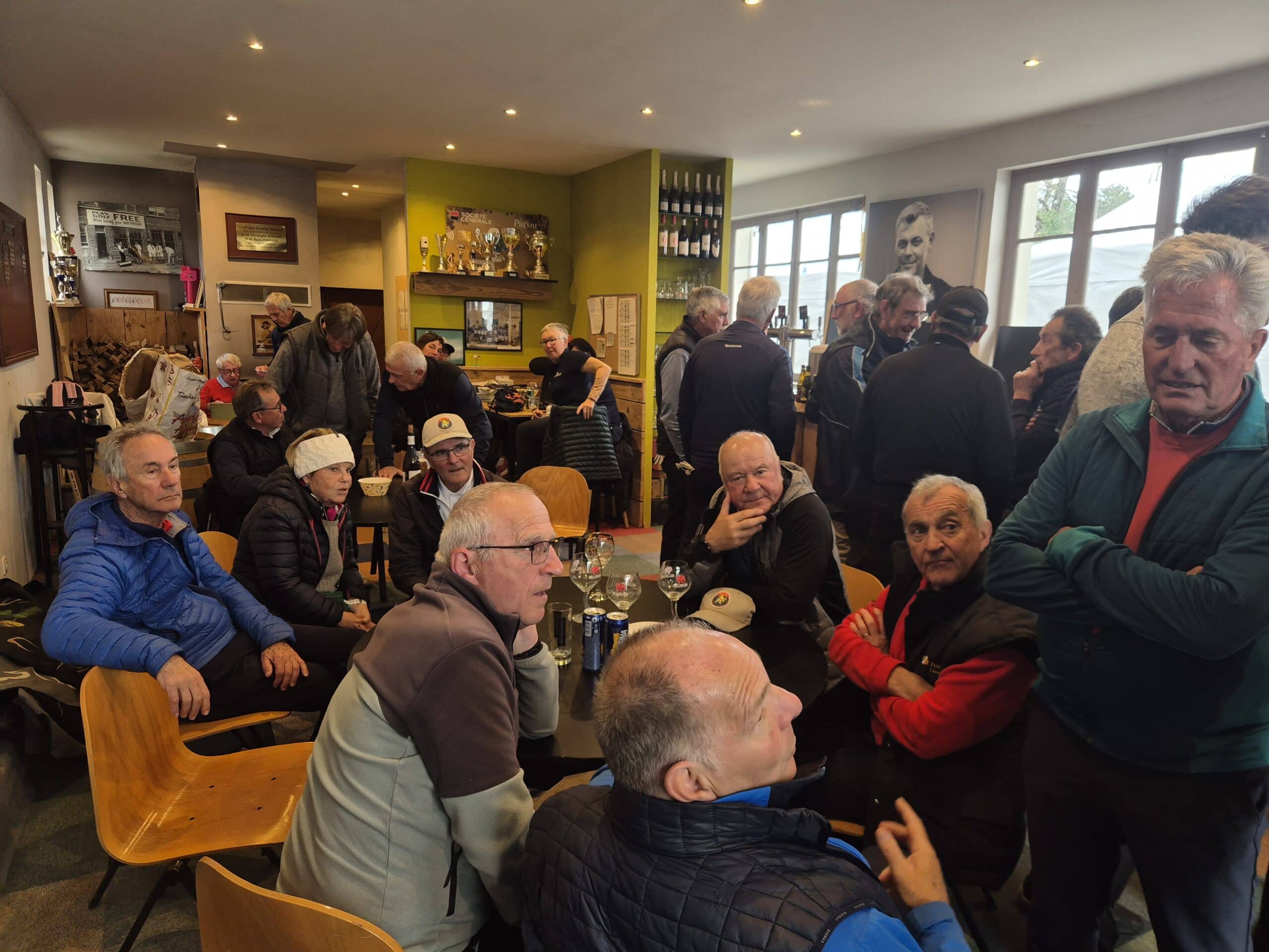 Un groupe de personnes se retrouve dans un café convivial, où l'ambiance est animée. Des hommes et des femmes de différents âges sont assis autour de tables, échangeant des rires et des discussions. L'intérieur du café est décoré avec des trophées et des souvenirs sportifs, créant une atmosphère chaleureuse. Des boissons sont visibles sur la table, et les participants semblent profiter d'un moment de camaraderie. Ce lieu est idéal pour les rencontres sociales et les événements communaut