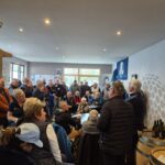 Groupe de personnes rassemblées dans un espace convivial, probablement lors d'un événement lié au golf. Des participants attentifs écoutent un orateur devant une table, tandis que d'autres sont assis sur des chaises. L'ambiance est accueillante, avec des décorations murales et des tonneaux en bois visibles. L'image illustre un moment de partage et d'échange au sein d'une communauté passionnée par le golf.