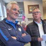 Deux hommes discutent dans un cadre convivial, probablement lors d'un événement sportif ou d'une réunion. L'un est vêtu d'un survêtement de sport avec un logo sur la poitrine, tandis que l'autre porte une veste et tient des documents dans ses mains. En arrière-plan, des membres d'un groupe semblent participer à l'activité, ajoutant à l'atmosphère animée de la scène. Ce moment illustre l'engagement communautaire et l'esprit d'équipe au sein d'une association