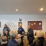 Réunion conviviale dans un club de golf, avec des participants attentifs écoutant deux intervenants devant un bar à vin. L'événement se déroule dans une salle lumineuse, décorée de manière moderne, et met en avant l'esprit communautaire du golf. Les participants, vêtus de tenues décontractées, semblent engagés dans des discussions animées, renforçant les liens entre les membres du club.