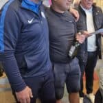 Trois hommes se tiennent côte à côte dans une ambiance conviviale, probablement lors d'un événement social. L'un d'eux, avec des cheveux gris, porte un vêtement de sport bleu et un sourire amical. Le deuxième homme, au centre, affiche une expression sérieuse tout en tenant une bouteille de vin. Le troisième homme, à droite, porte des lunettes et un gilet noir, regardant un document. En arrière-plan, on aperçoit des barriques et d'autres