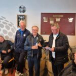 Cérémonie de remise de prix dans un club de golf, avec plusieurs hommes réunis autour d'un bar. L'un des hommes, tenant une bouteille, reçoit un prix tandis que deux autres le regardent, l'un lisant un discours. L'ambiance est conviviale et festive, avec des membres du club assis en arrière-plan. Décor intérieur moderne avec des éléments de golf visibles.