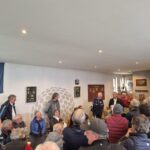 Réunion de golfeurs dans un club, avec plusieurs hommes attentifs à un discours. En arrière-plan, des trophées et des souvenirs de golf sont exposés. Une bannière de la fédération française de golf est visible, indiquant l'importance de l'événement. L'atmosphère est conviviale et engageante, mettant en avant la passion pour le golf et le partage d'expériences entre les membres du club.