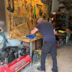 Atelier de bricolage bien équipé avec un mur d'outils variés accrochés sur une planche en bois. Un homme travaille sur un établi, utilisant des outils pour des projets de réparation ou de construction. Un compresseur d'air rouge est visible au premier plan, tandis que des étagères en arrière-plan contiennent divers matériaux et équipements. L'éclairage est adéquat, créant un environnement de travail fonctionnel et organisé.