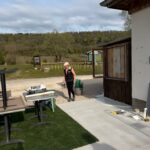 Une femme en tenue décontractée, portant une casquette, se trouve à l'extérieur d'un bâtiment en bois, en train de porter un seau. L'environnement est verdoyant, avec des collines en arrière-plan et des équipements de jardinage visibles à proximité. Le sol est recouvert de gazon synthétique et des tables sont disposées sur une surface en béton. Cette image illustre une activité de préparation ou d'aménagement dans un espace extérieur, propice aux événements ou